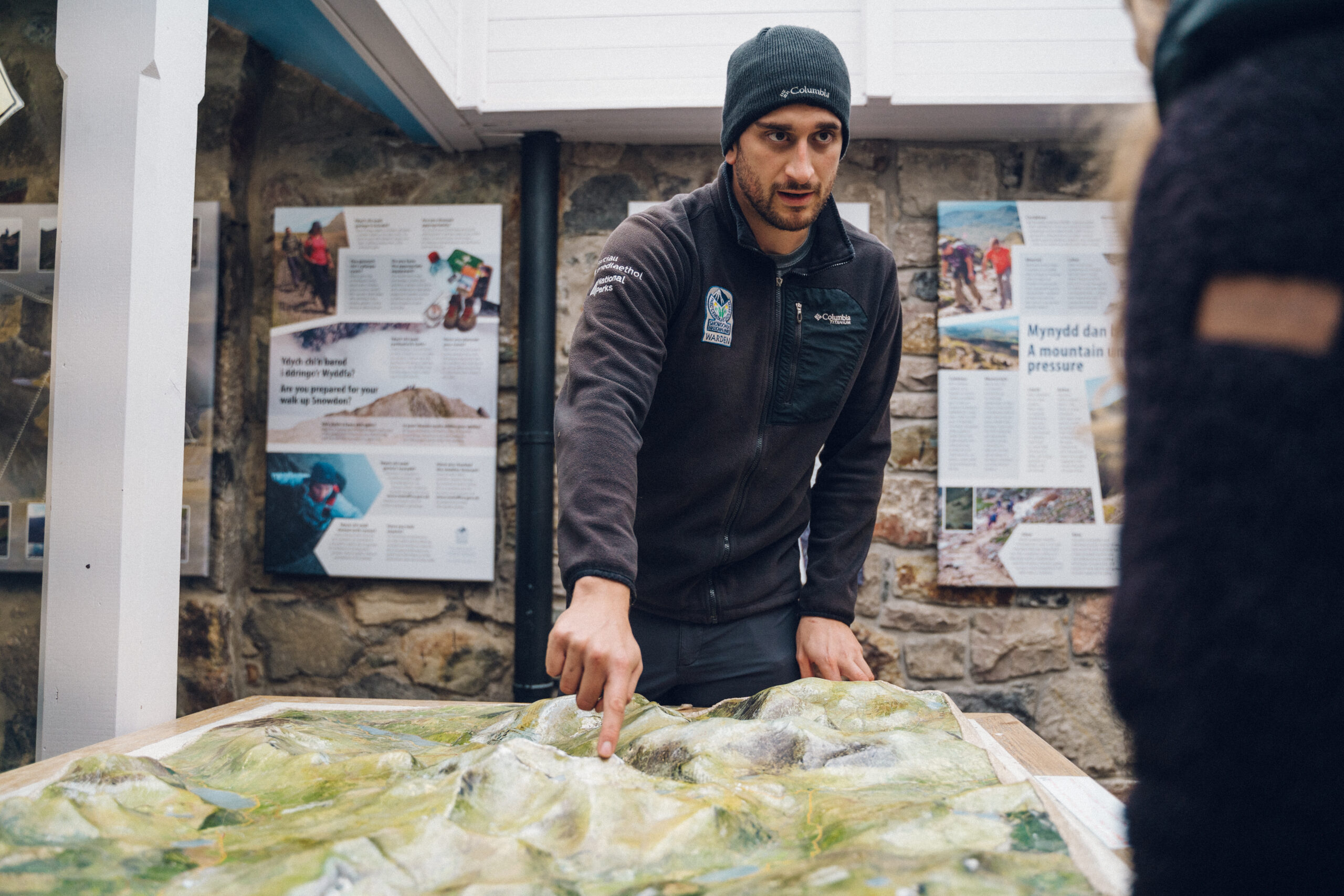 Warden y Parc Cenedlaethol yn gwisgo het wlanog a siaced awyr agored, yn pwyntio at fap dan do, gyda phaneli gwybodaeth a wal garreg yn y cefndir. A National Park Warden in a beanie and outdoor jacket is pointing at a raised map display indoors, with informational panels and a stone wall in the background.