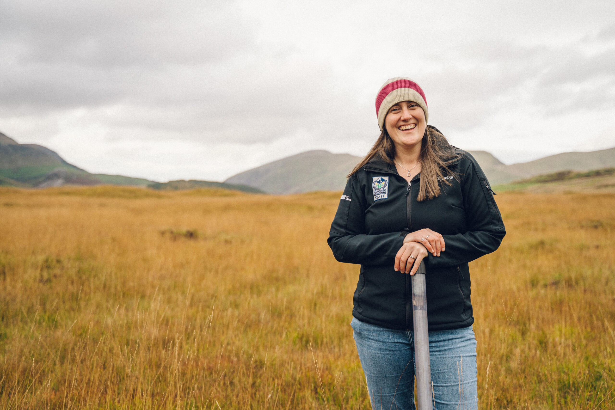 Merch yn gwenu, yn gwisgo het wlanog a siaced awyr agored Parc Eryri, yn sefyll mewn cae o laswellt tal, gyda mynyddoedd yn y cefndir. A smiling woman in a beanie and outdoor jacket stands in a field of tall grass, holding onto a tool, with rolling hills and a cloudy sky in the background.